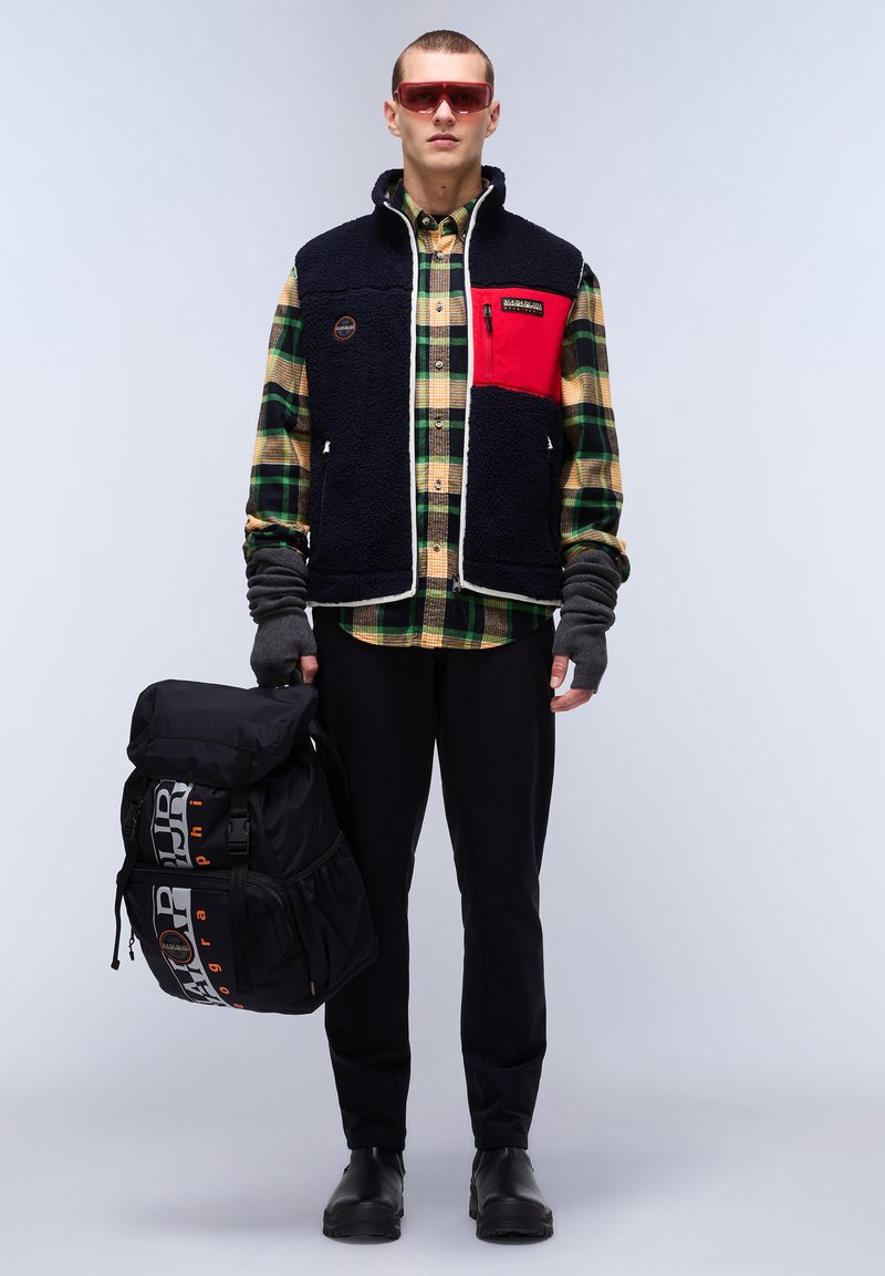 Gilet en polaire marine avec une accent rouge, associé à une chemise à carreaux verte et un pantalon noir. Le sac à dos noir présente des lettres grises et un rabat.