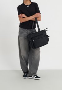 Black nylon tote bag with adjustable straps, front zip pocket, side pockets, and a smooth texture. Paired with gray wide-leg jeans and black shoes.