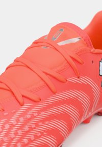 Botín de fútbol rojo brillante con parte superior texturizada, que presenta patrones angulares blancos y cordones a juego, diseñado para rendimiento y agarre.