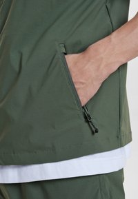 Armádně zelená bunda se zipovou kapsou, s hladkým povrchem a bílým vrstveným lemem viditelným na spodní části.