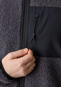 Ciemnoszary polar z czarną częścią z materiału. Posiada kieszeń zapinaną na zamek oraz logo "SWEDEMOUNT", ręka trzymająca uchwyt zamka.