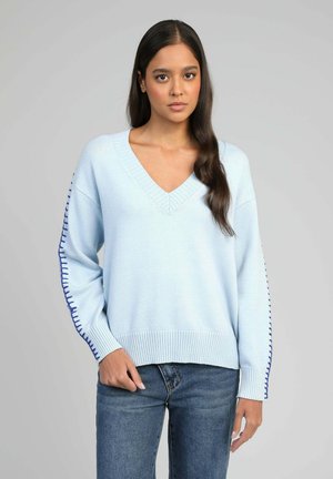Femme aux longs cheveux foncés portant un pull léger bleu clair à col en V avec des coutures bleu foncé sur les manches et un jean bleu, se tenant devant un fond gris.