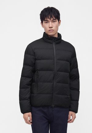 Jeune homme aux cheveux courts noirs portant une veste matelassée noire avec fermeture éclair et un pantalon foncé, debout devant un fond uni clair.