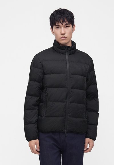 Jeune homme aux cheveux courts noirs portant une veste matelassée noire avec fermeture éclair et un pantalon foncé, debout devant un fond uni clair.