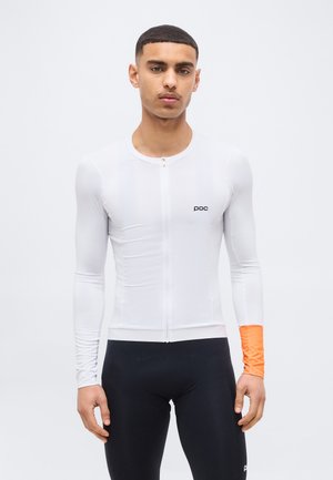 Jonge man die een witte fietstrui met lange mouwen en rits draagt, met een oranje linker manchet en zwarte fietsbroek, staand tegen een witte achtergrond.