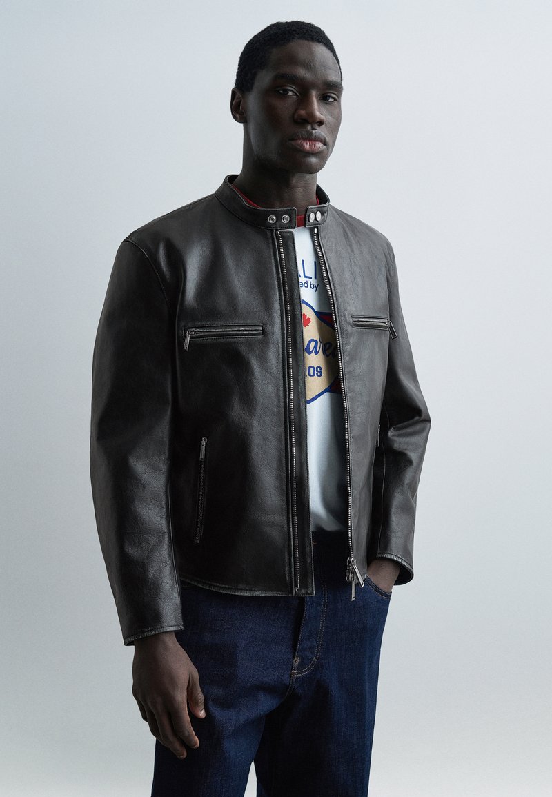 Νέος άνδρας που φοράει μαύρο δερμάτινο μπουφάν πάνω από ένα λευκό μπλουζάκι με τύπωμα και σκούρο μπλε τζιν, στέκεται με το ένα χέρι στην τσέπη.