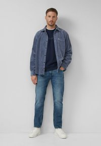 Uomo che indossa una camicia di velluto blu sopra un maglione navy, jeans blu e sneakers bianche, in piedi su uno sfondo grigio chiaro uniforme.