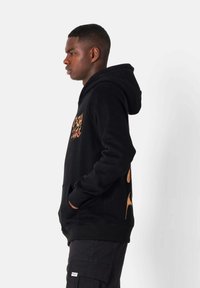 Sudadera negra con bolsillo delantero, capucha ajustable y gráfico de llamas naranjas en el lateral. Tejido texturizado con puños y dobladillo acanalados.