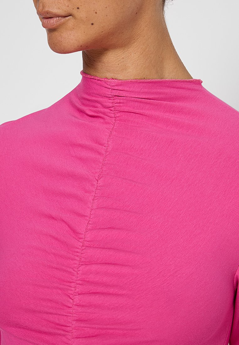 Rosa High-Neck-Top aus weichem Material, mit gerafften Details entlang der Rückennaht und einer glatten, dehnbaren Textur.