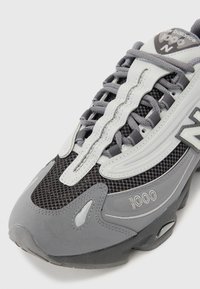 Sneaker atletico grigio e grigio chiaro con pannelli in rete, sovrapposizioni testurizzate e suola in gomma; presenta il marchio "1000" sul lato.