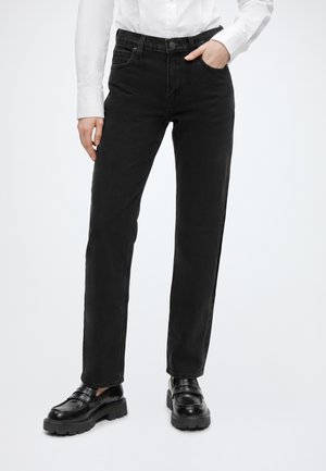 Personne portant un jean noir à jambes droites, des mocassins noirs épais et une chemise blanche à manches longues, une main dans la poche.