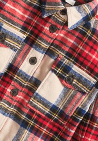 Chemise en flanelle à carreaux rouge, bleu et jaune avec une base beige ; comporte deux poches poitrine et des boutons noirs ; tissu doux et texturé.