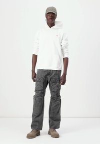 Hanorac alb cu buzunar frontal tip cangur, pantaloni cargo gri cu multiple buzunare, cizme bej și o șapcă gri deschis. Design minimalist.
