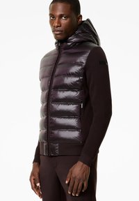 Gilet imbottito con una parte frontale lucida di colore viola scuro e lati in maglia a coste marrone scuro. Presenta un cappuccio e una chiusura con zip. Branding discreto sulla manica.