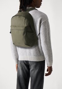 Mochila verde oliva con correas ajustables, bolsillo frontal con cremallera y tela suave. Llevada con un suéter de punto gris claro y jeans oscuros.