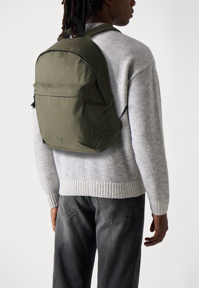 Mochila verde oliva con correas ajustables, bolsillo frontal con cremallera y tela suave. Llevada con un suéter de punto gris claro y jeans oscuros.