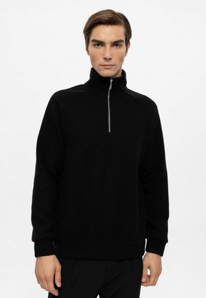 Pull-over noir avec col montant, fermeture éclair mi-longue à l'avant, tissu texturé, poignets et ourlet côtelés, porté avec un pantalon noir.