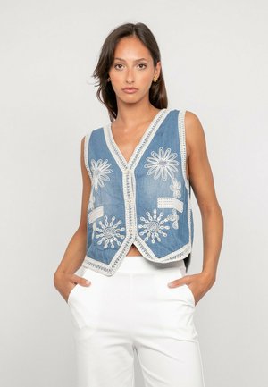 Femme portant un gilet sans manches bleu avec des motifs floraux crochetés blancs et un pantalon blanc, debout les mains dans les poches devant un fond uni.