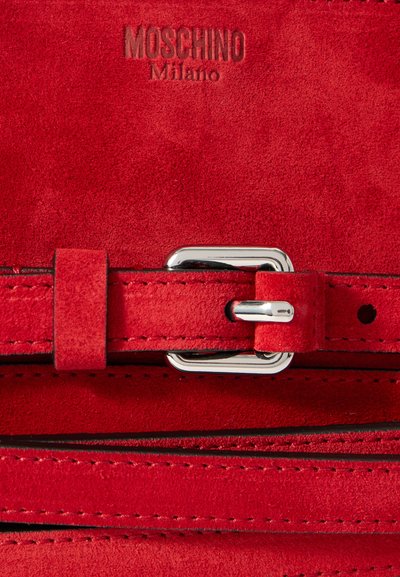 Detalj av röd mockasäck med en silvermetallspänne och sömnad. Den präglade "Moschino Milano"-logotypen är synlig.