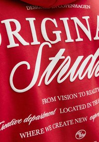 Roter Kapuzenpullover mit großem weißem Text. Auffällige Schriftzüge mit der Aufschrift "ORIGINAL STUDIOS" sowie kleinerem Text, der die Kreativabteilung und den Standort beschreibt.