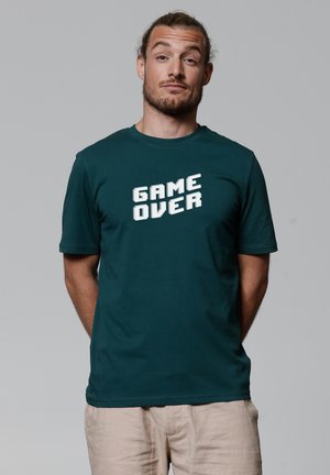 T-shirt à manches courtes teal en coton, avec un grand graphique blanc "GAME OVER" en police pixelisée sur le devant.