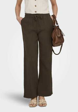 Žena odjevena u smeđe teksturirane hlače širokih nogavica s vezicama u struku, bež pletene sandale, kremasti bezrukavi gornji dio s gumbima, držeći smeđu kožnu torbu.