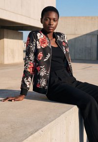 Junge Frau in schwarzer, geblümter Jacke und schwarzer Hose sitzt auf einer Betonkante in urbanem Außenbereich unter blauem Himmel.