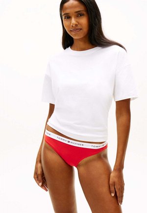 Frau trägt ein weißes cropped T-Shirt und rote Tommy Hilfiger Unterwäsche mit Logobund, steht vor einem einfarbig weißen Hintergrund.