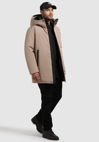 Parka acolchada beige con capucha, cremallera negra y acentos, llevada sobre un suéter negro y pantalones, combinada con botas negras.