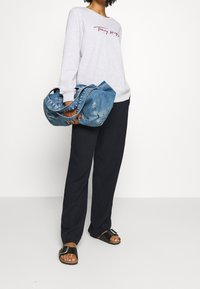 Šedá mikina s červeným logem, volné černé kalhoty a černé sandály. Drží měkkou modrou džínovou tašku se dvěma uchy.