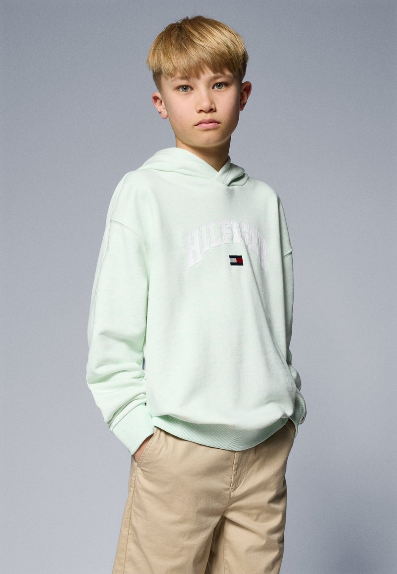 Svetlo mint zelena kapuca z sprednjim žepom in belim logotipom "HILFIGER". Ujemite z bež kratkimi hlačami. Mehka tekstura in sproščen kroj.