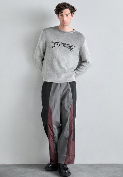 Pull en maille gris avec un texte graphique noir, associé à un pantalon ample gris et bordeaux à panneaux divisés. Chaussures : baskets noires épaisses.