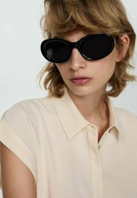 Zwarte oversized zonnebril met een glanzende afwerking, gecombineerd met een lichtbeige overhemd met kraag. Het overhemd heeft opgerolde mouwen en een soepele textuur.