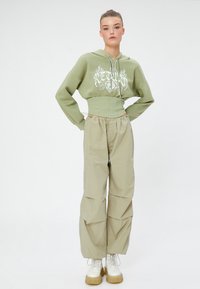 Pantalones caqui de cintura alta combinados con un hoodie corto de color oliva con texto gráfico en blanco. Botas blancas de suela gruesa completan el conjunto.