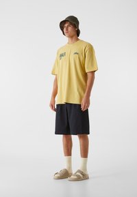 Gul kortermet t-skjorte med grafisk trykk, kombinert med mørk marineblå shorts. Tilbehør inkludert en grønn strikket bøttehatt og beigefargede sandaler.