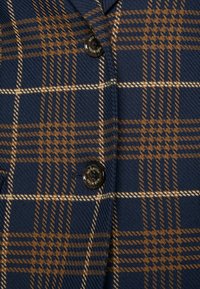 Tissu à carreaux bleu marine et marron avec une texture diagonale présentant des boutons noirs étiquetés "MOS MOSH" sur un vêtement.