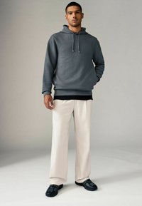 Grijze textuur hoodie met trekkoord, gelaagd over een zwart shirt. Gepaard met lichtbeige wijde broek en zwarte sneakers.