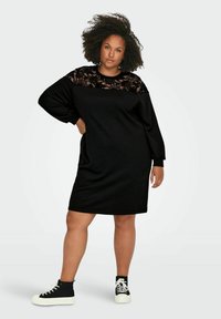 Vestido negro con un panel superior de encaje, mangas largas y corte holgado. Combinado con zapatillas negras y blancas y un peinado texturizado.