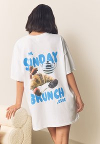 Camiseta blanca de gran tamaño con un gráfico colorido de un croissant, fresas y una tetera con el texto "THE SUNDAY BRUNCH .club".