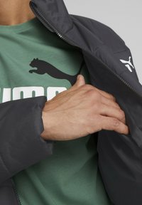 Giacca nera, isolante, con colletto alto, indossata sopra una maglietta verde con il logo Puma nero. Mano che toca l'area del petto della giacca.