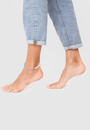 Perlebesat ankelkæde med blå kædelinker og hvide perler, båret på en bar ankel, sammen med opfoldede lyse denimjeans.