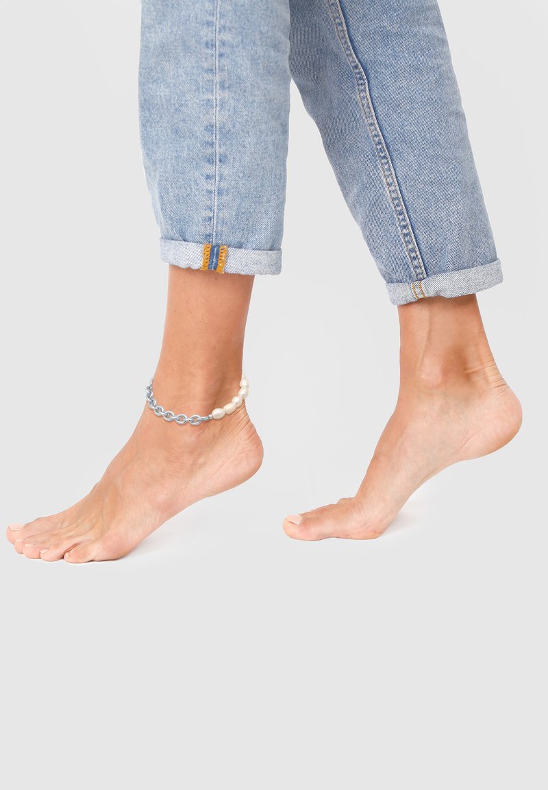 Perlebesat ankelkæde med blå kædelinker og hvide perler, båret på en bar ankel, sammen med opfoldede lyse denimjeans.