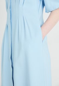 Robe bleu clair en tissu lisse, avec de courtes manches bouffantes, des plis verticaux et des poches latérales. Coupe ajustée avec des lignes épurées.
