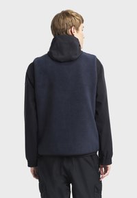 Mornarska flis prsluk s visokim ovratnikom, s mekim teksturom. Prsluk nema vidljivih kopčanja i nosi se preko crne majice s dugim rukavima.