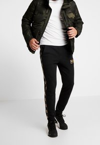 Mężczyzna w czarnych dresowych spodniach Puma z bocznym paskiem z logo, czarnych sneakersach, białej koszulce i ciemnozielonej kurtce w camo, stojący na białej podłodze.