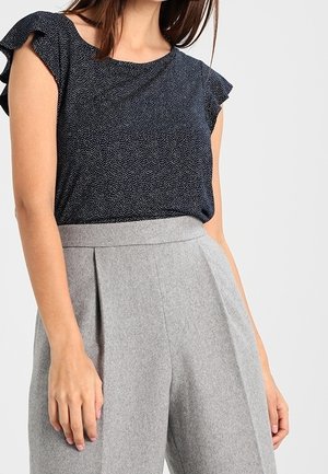 Femme portant un chemisier sans manches bleu marine à pois avec des épaules à volants et un pantalon plissé gris taille haute.