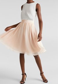 Lichtroze, knielange rok met een vloeiende silhouet en elastische tailleband, gecombineerd met een witte mouwloze top. Zwarte sandalen met bandjes.
