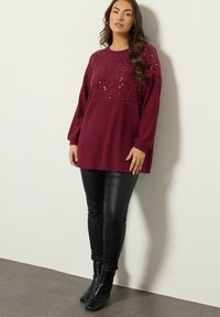 Bordowy sweter dzianinowy z cekinowymi ozdobami w górnej części, zestawiony z czarnymi legginsami z ekoskóry i botkami. 