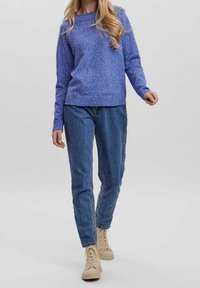 Frau, die an einem einfarbigen hellen Hintergrund geht und einen blauen Pullover, blaue Jeans und beigen High-Top-Sneakers trägt.