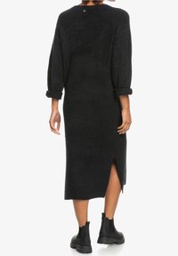 Langes schwarzes Strickkleid mit figurbetonter Silhouette, aufgekrempelten Ärmeln, Seitenschlitz und Knopfdetail am Rücken. Kombiniert mit schwarzen Knöchelstiefeln.